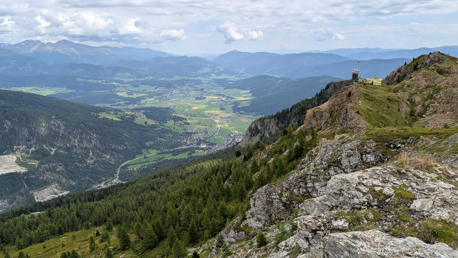 A wide view from the Speiereck down over a green valley floor of fields and villages, with a small tower on the rocky crest to the right.