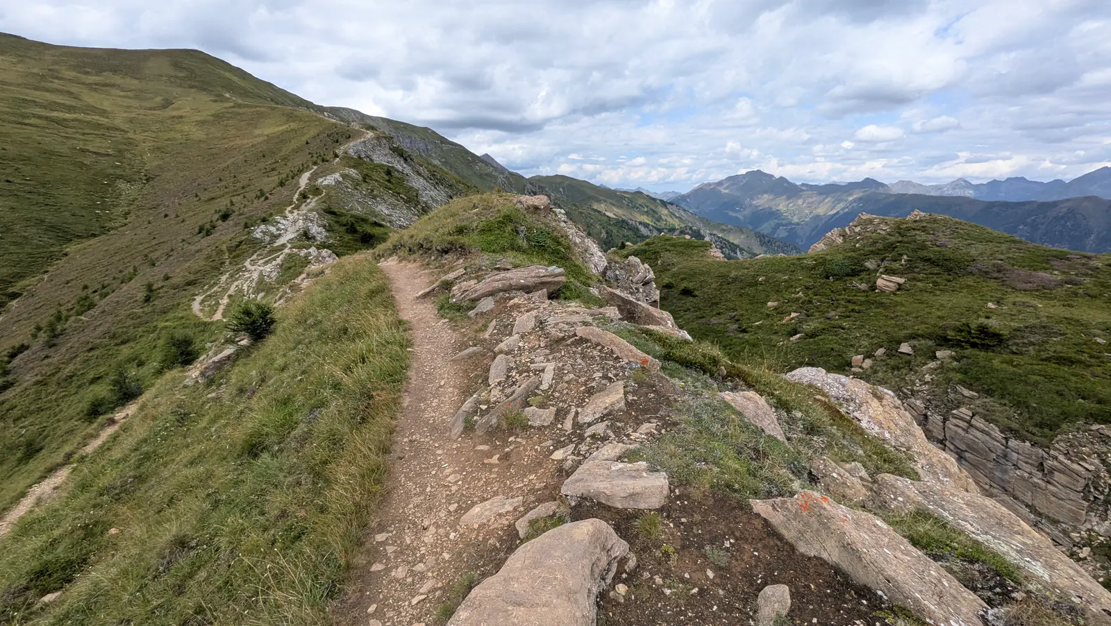 A narrow gravel path runs along a grassy ridge on the Speiereck between rocky outcrops, with ranges of mountains fading into the distance.