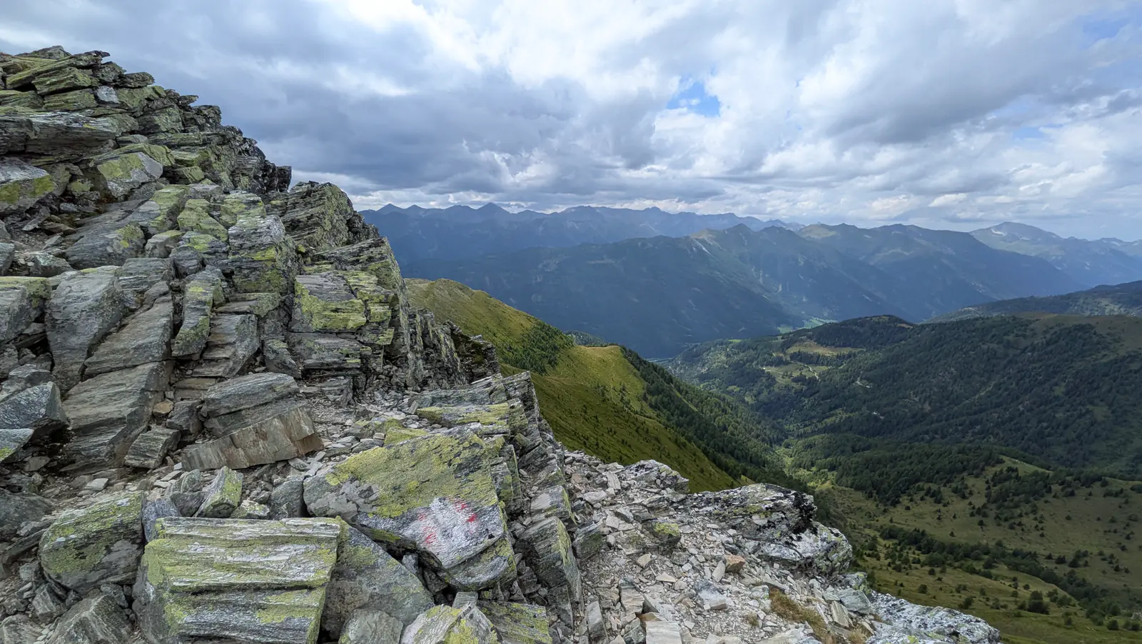 Layered slabs of lichen-covered rock on the Speiereck summit ridge carry a red and white painted waymark, above a green valley and distant peaks.