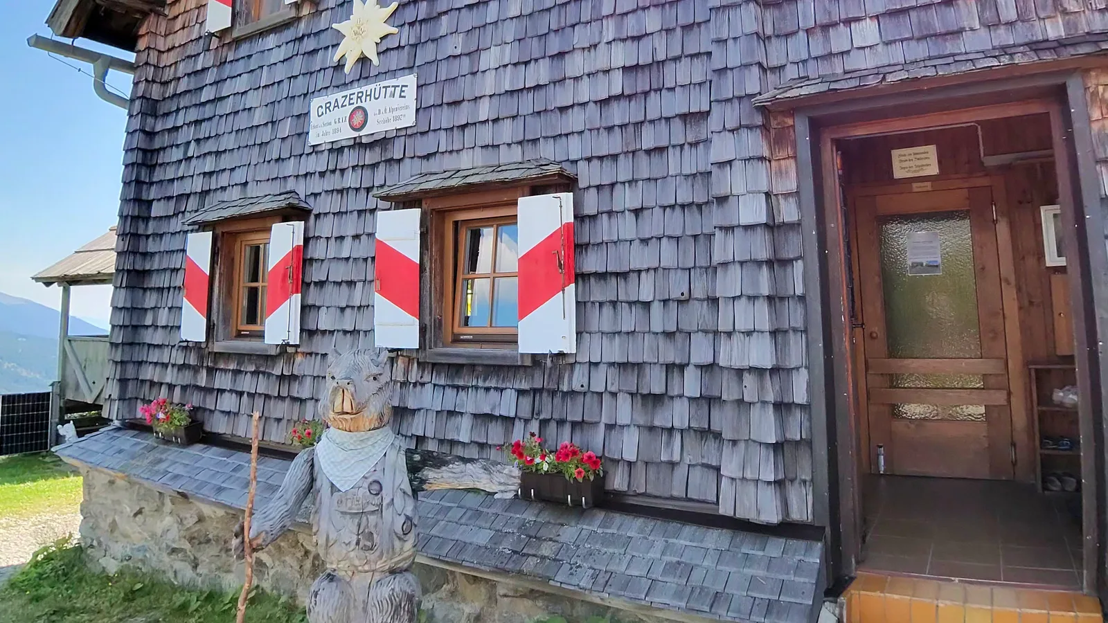 Shingled facade of the Grazer Hütte with red-white shutters, the hut sign, a carved marmot figure and the entrance door in summer.
