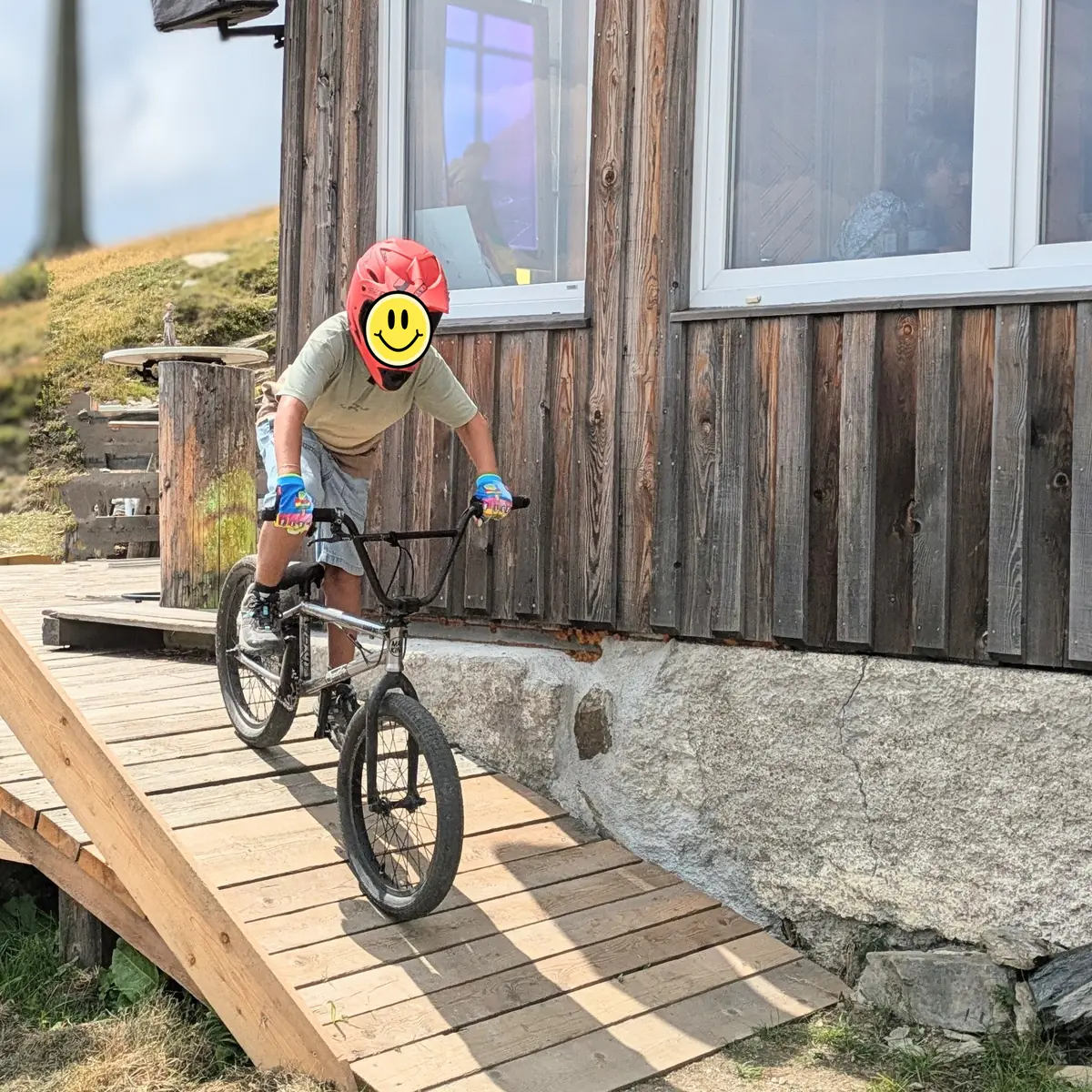 A young rider in a red helmet balances a BMX on a wooden ramp against the weathered larch wall of the Speiereckhütte.