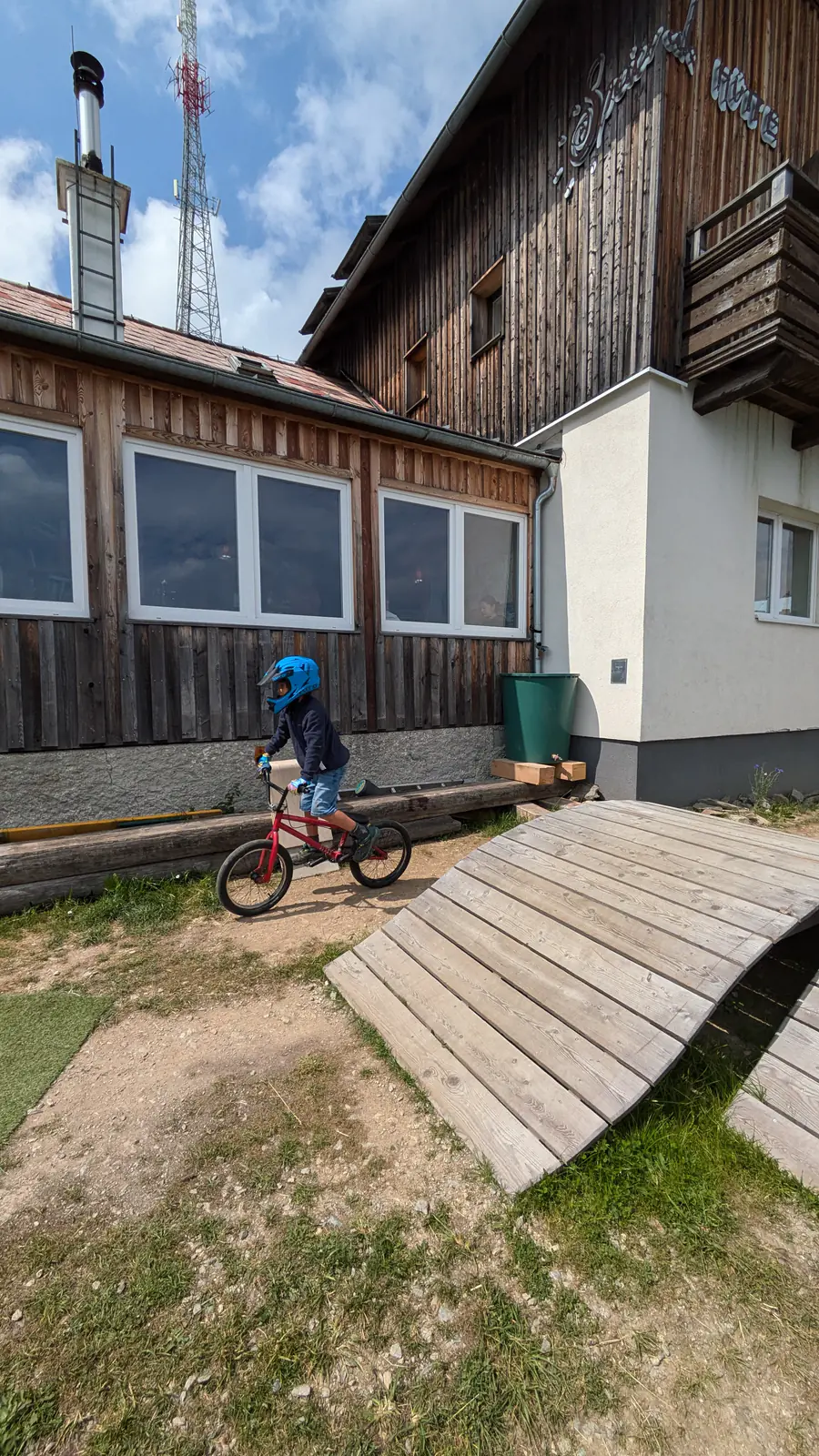 A child in a full-face helmet rides a small red BMX past a curved wooden ramp below the timber facade of the Speiereckhütte.