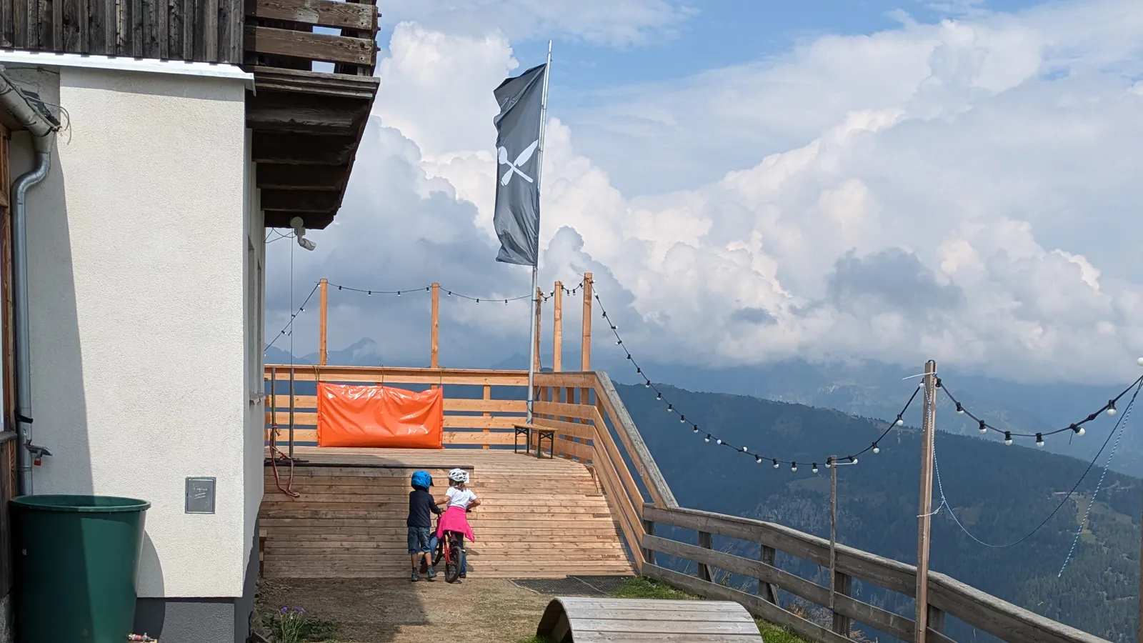The wooden sun terrace of the Speiereckhütte with a restaurant flag and string lights, looking out over forested slopes and towering summer clouds.